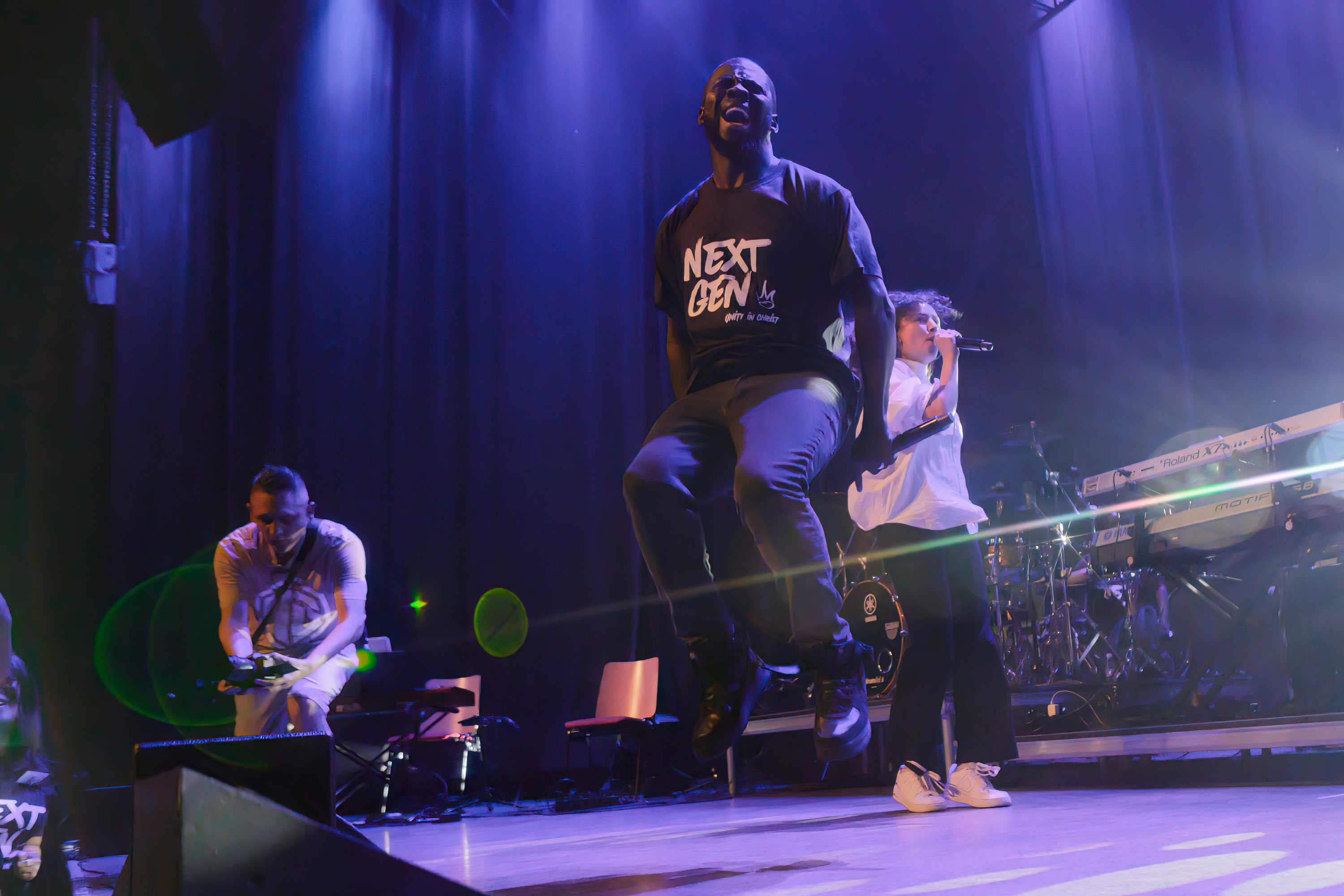 Godwin Arhin springt in een zwart Next Gen shirt hoog boven een paars verlicht podium, met band op de achtergrond