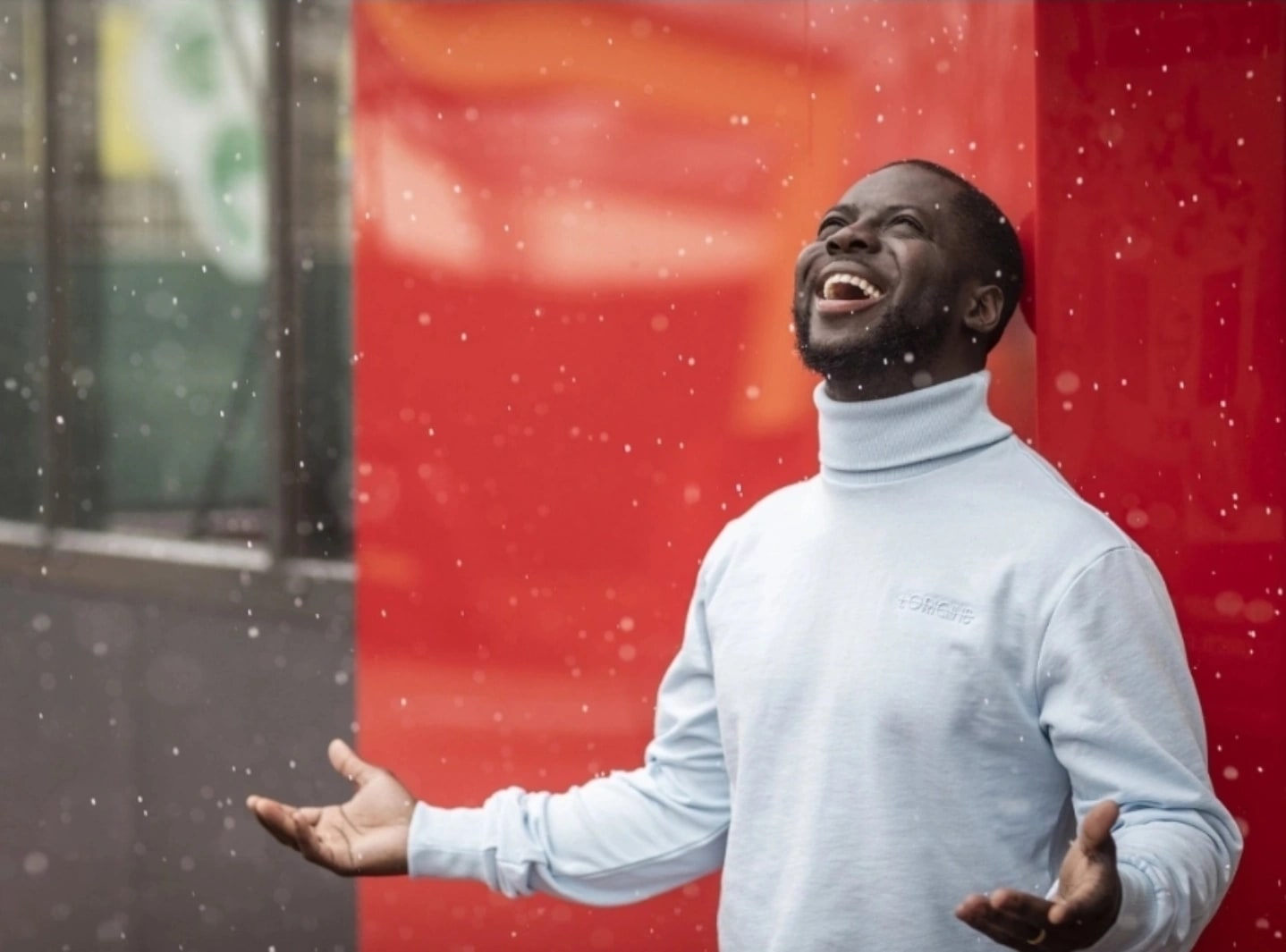 Godwin Arhin kijkt lachend omhoog met gespreide armen in de vallende sneeuw, in lichtblauwe coltrui tegen een felrode wand