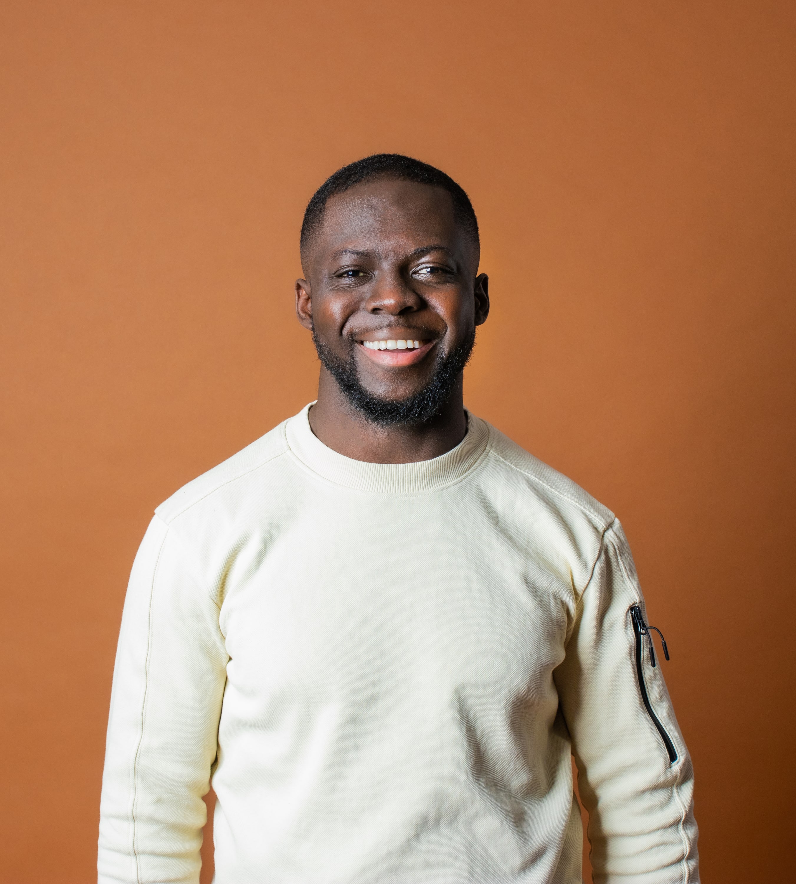Godwin Arhin lacht breed in een crèmekleurige trui tegen een warme oranjebruine studioachtergrond